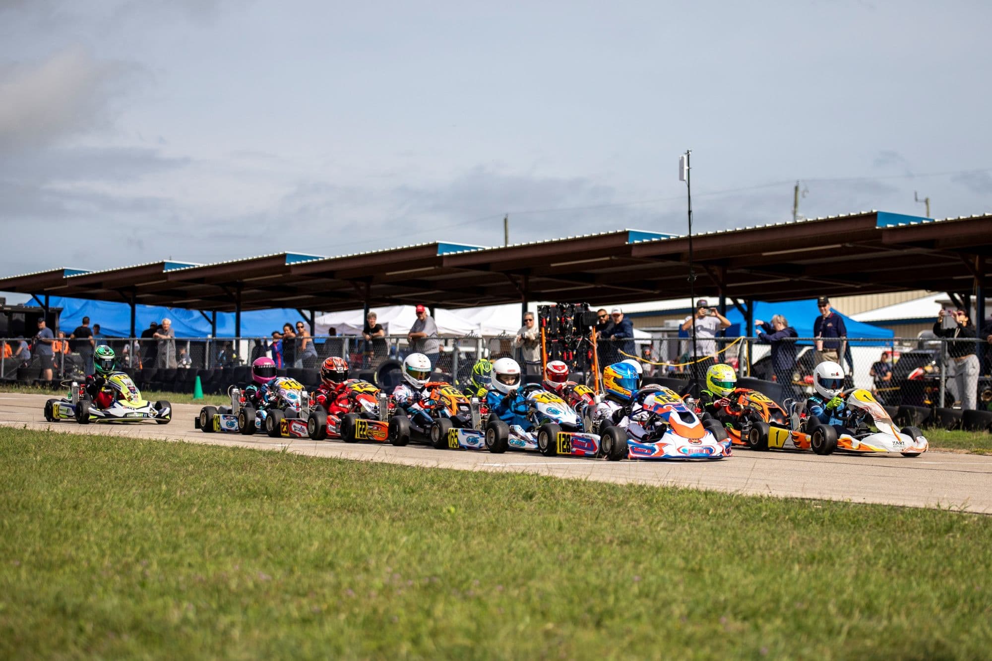 A full kart grid lined up under the spectator pavilion at KartMoto, under a gray sky.